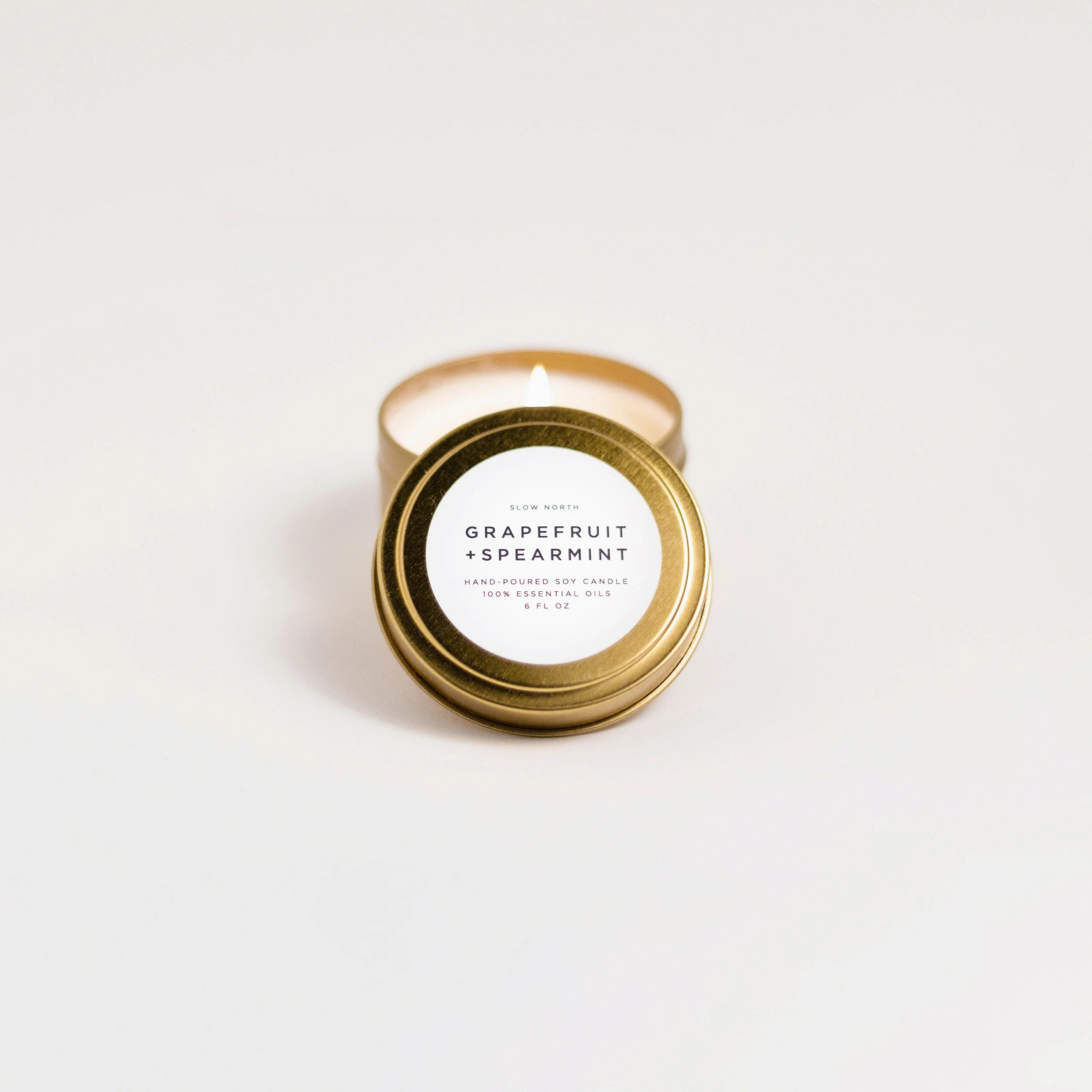 Lit grapefruit spearmint scented soy candle in gold travel tin on white surface