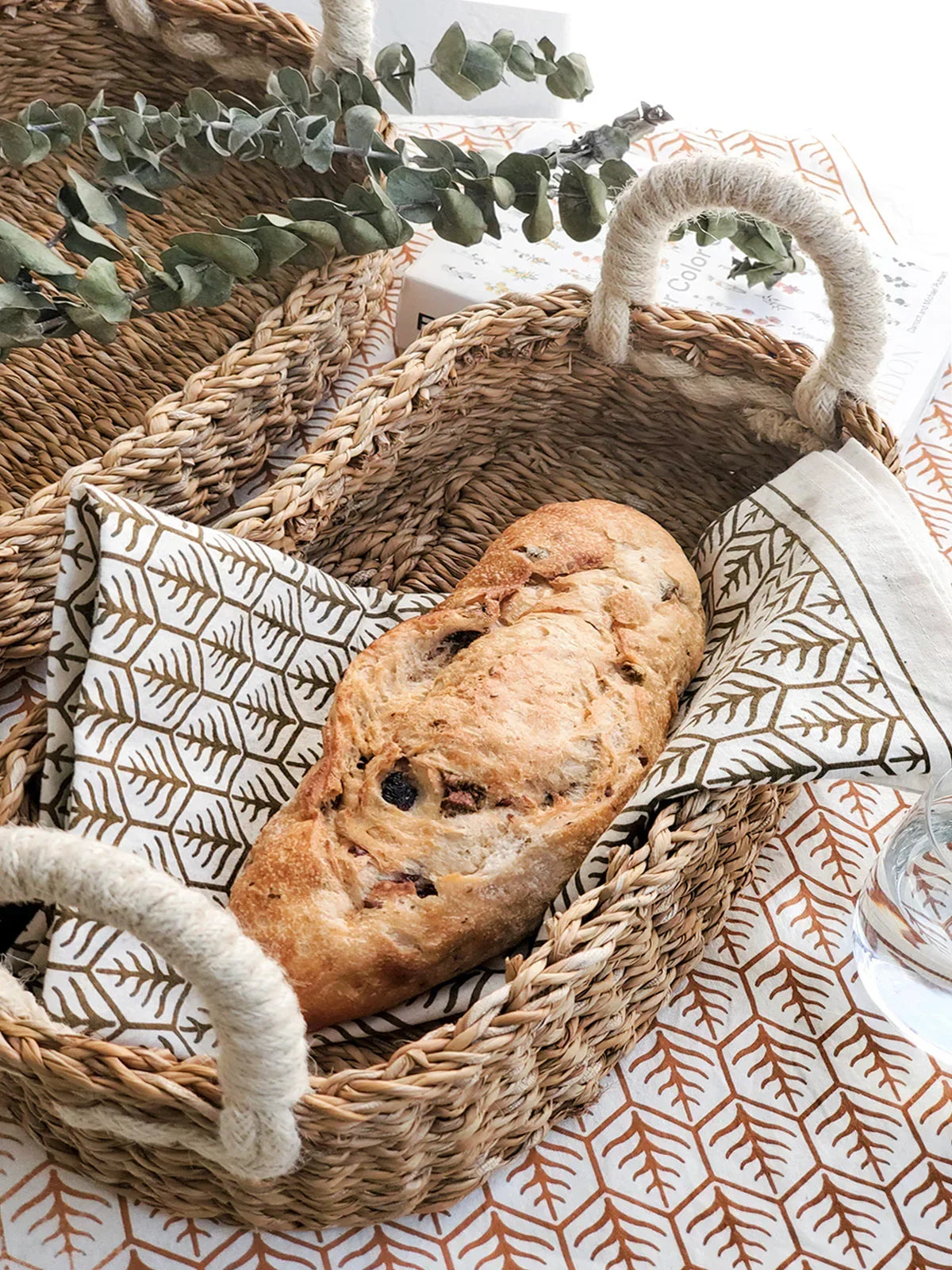 Oval woven bread basket with handles, holding a patterned cloth and a loaf of rustic bread