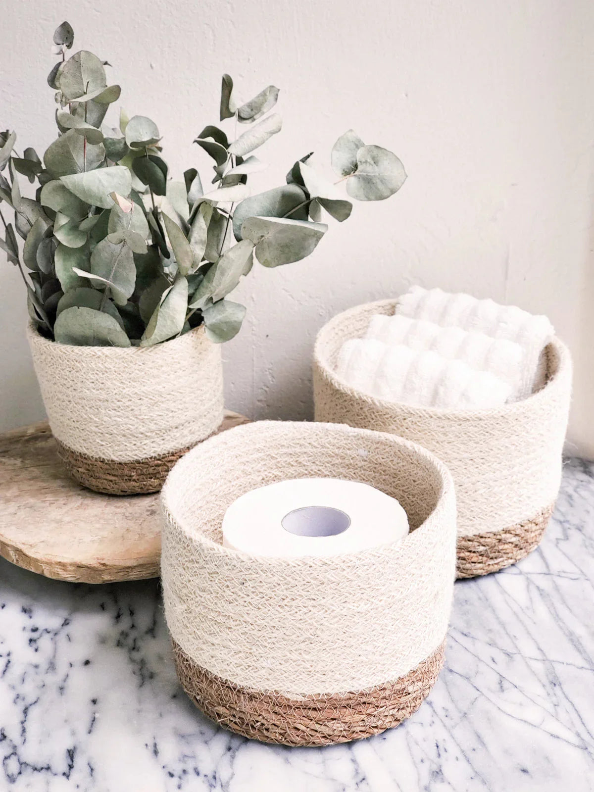 Set of three woven storage baskets on marble table with eucalyptus and towels